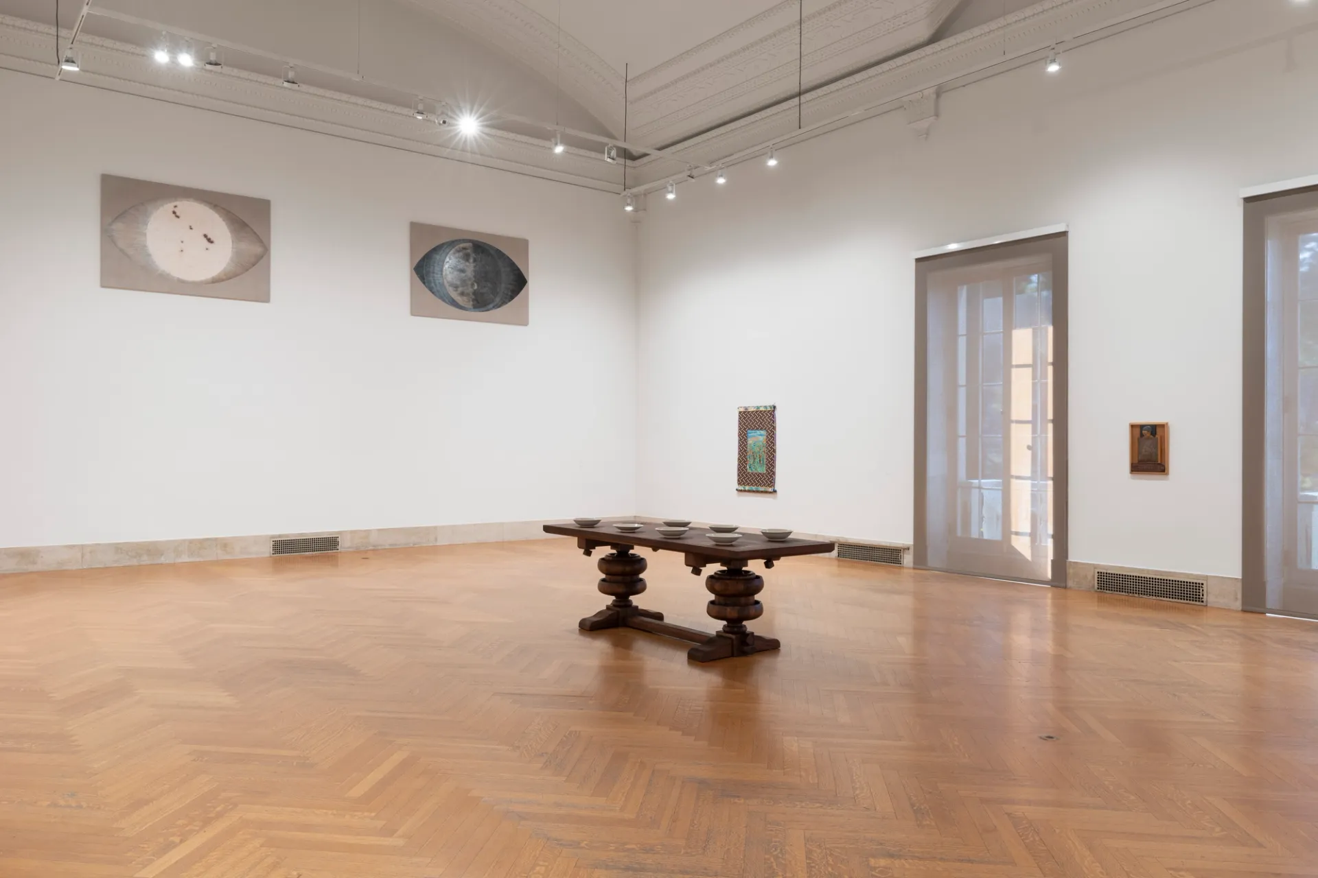 A carved wooden table supporting a group of small ceramic sculptures stands in the center of a space. To the left, two artworks are positioned high on the wall. To the right, two artworks are on the wall on either side of a large window.