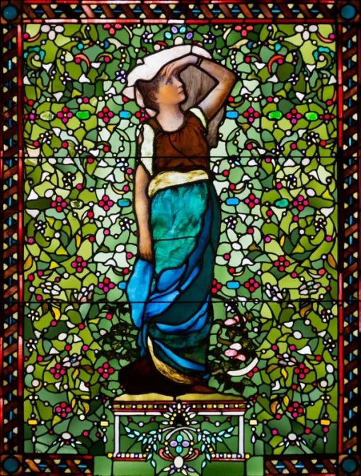 Vibrant stained glass window with a woman shown standing at center and looking upwards, with one arm held to her forehead. She is surrounded by abstract forms that evoke leaves and flowers and a decorative frame.