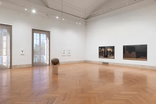 To the left, floor-to-ceiling windows with framed artwork on the walls. On the wall to the right, two large horizontal paintings, and in the center of the floor, sits a sculpture of a large wooden orb atop a wooden cube 