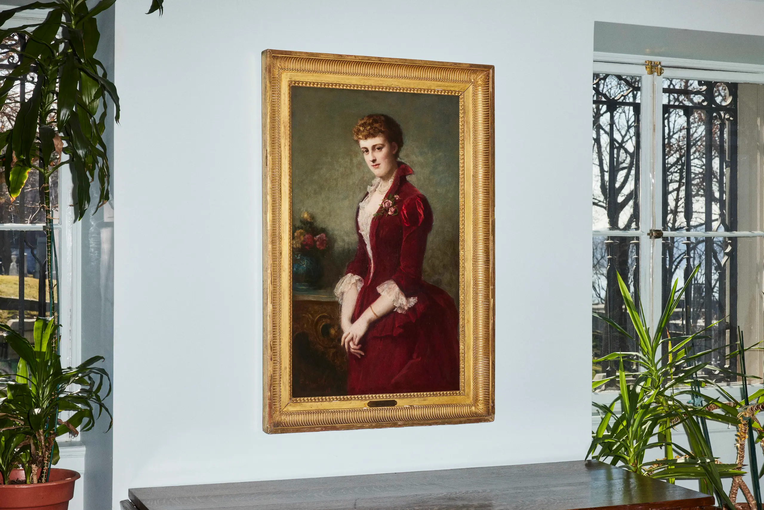 A painting of Edith Wharton hanging on a wall beside plants and two windows.