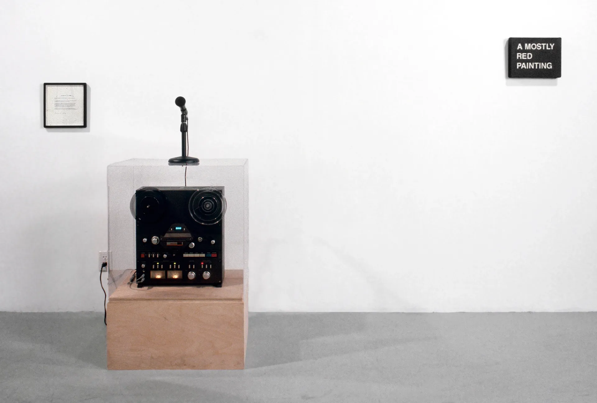 A reel-to-reel tape recorder sits on a wooden plinth, covered by an acrylic box with a microphone on top. A black rectangle hangs on the wall to its right with white text saying "a mostly red painting."