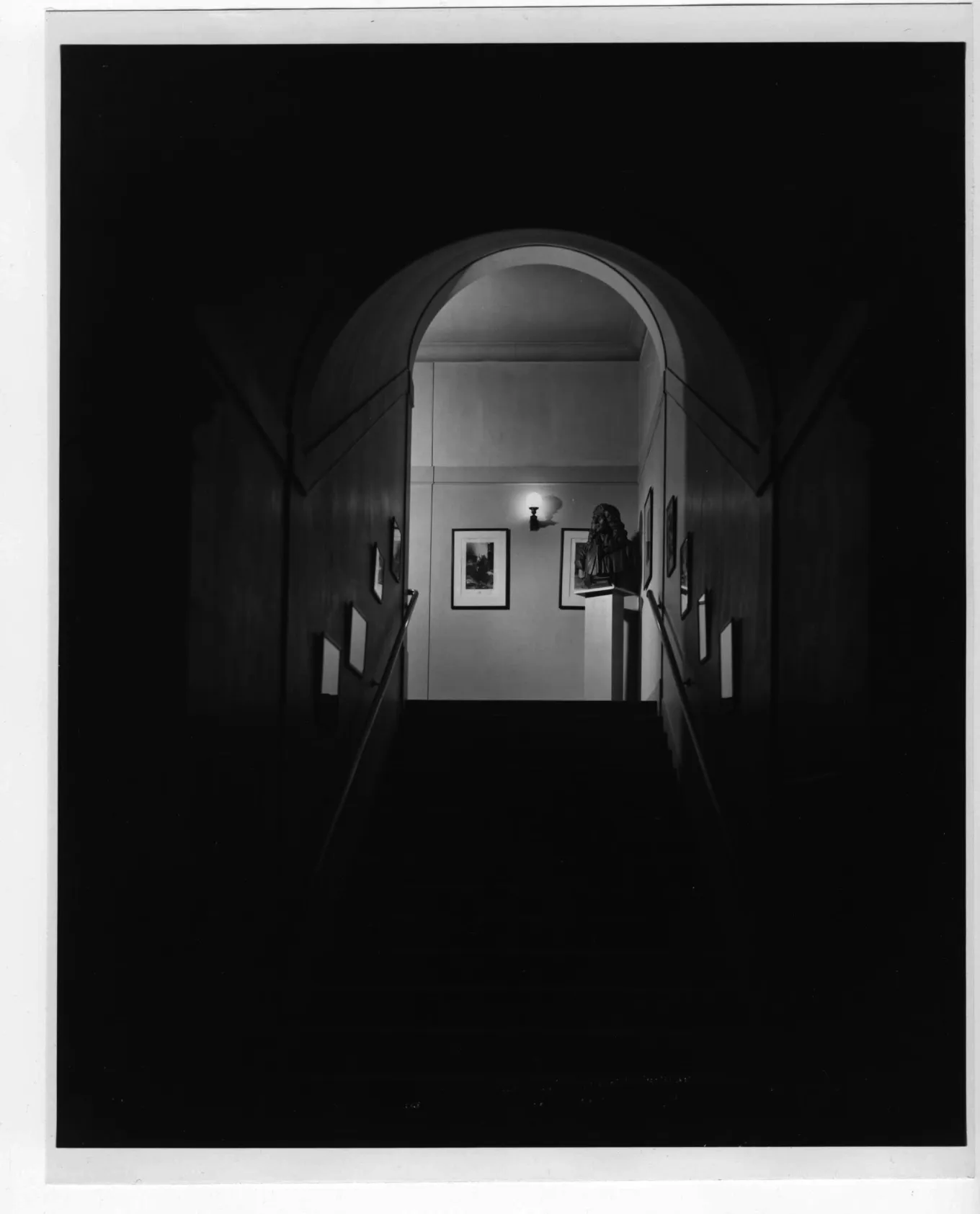 A black and white photograph of a dark stairwell that leads up to the arched doorway of a brightly lit room. Framed objects hang along the stairwell walls and within the room atop the stairs.