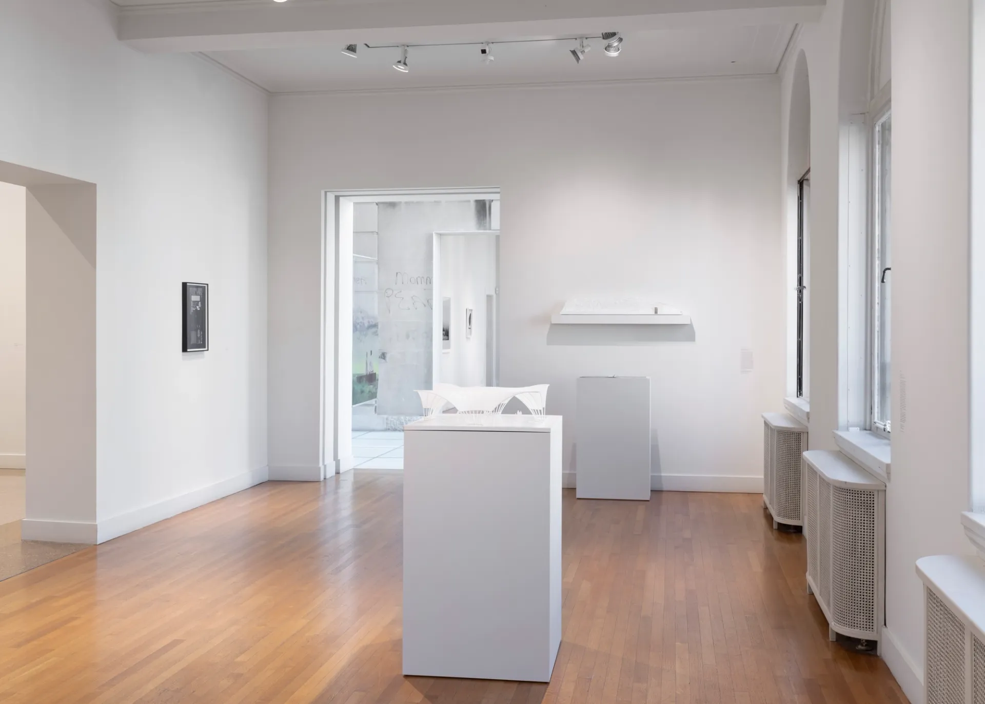 White architectural models sit positioned inside a room and on a wall to the right. To the left, a single framed artwork is on the wall. A gallery ahead on the left contains more artworks.