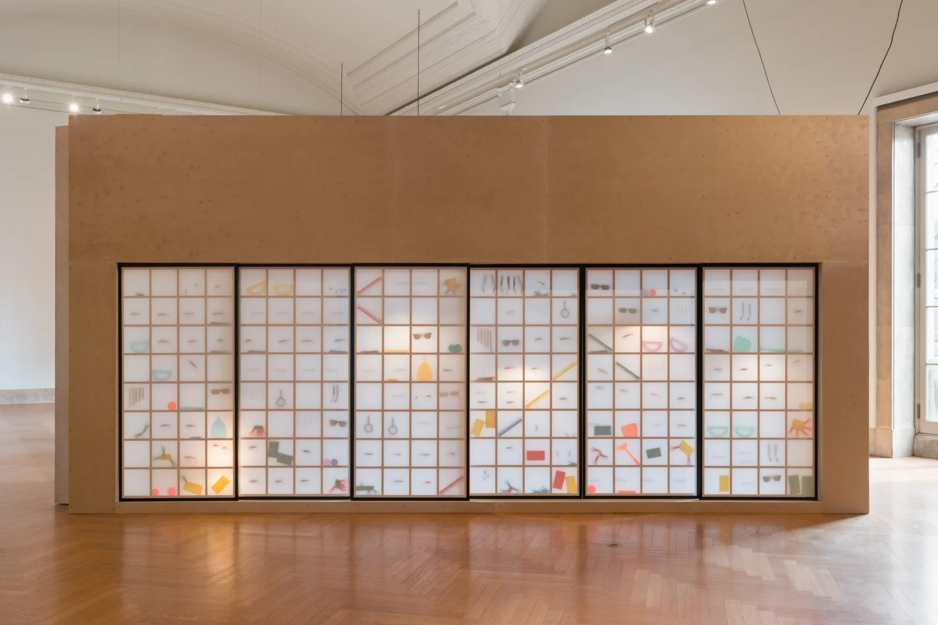 An art installation with a row of siding paper doors with objects and a gridded wooden frame visible inside. 