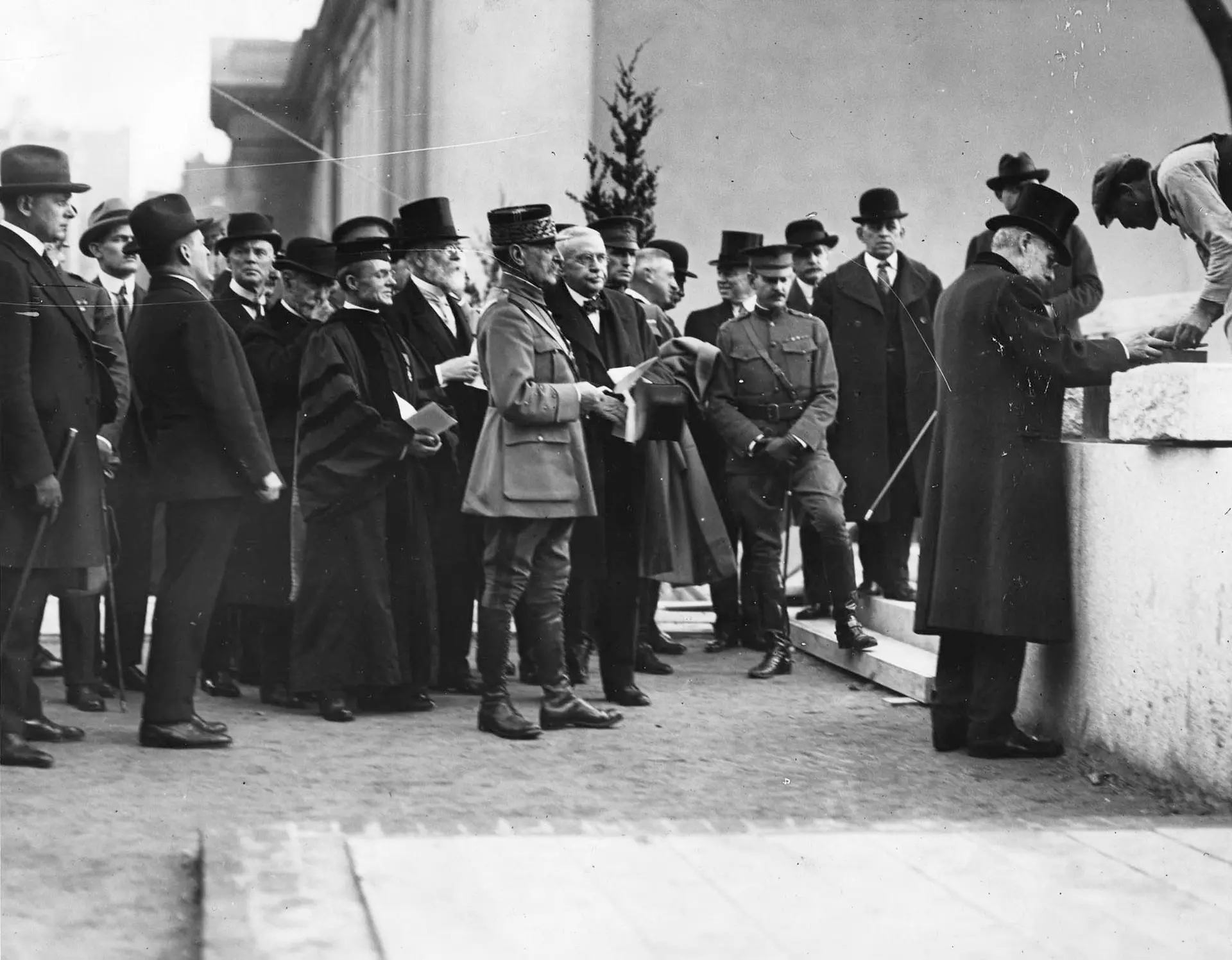 A black and white photograph of a group of men wearing suits and top hats looking towards two men setting a stone atop a waist high wall.