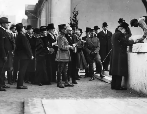 A black and white photograph of a group of men wearing suits and top hats looking towards two men setting a stone atop a waist high wall.