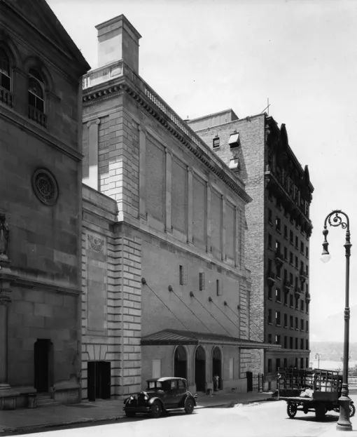 A black and white photograph of the American Academy of Arts and Letters building, set in between two other buildings made with darker stone. Two 1920s-era cars are parked in the street in front of the building. Behind the buildings is the Hudson River and surrounding hills in the distance.
