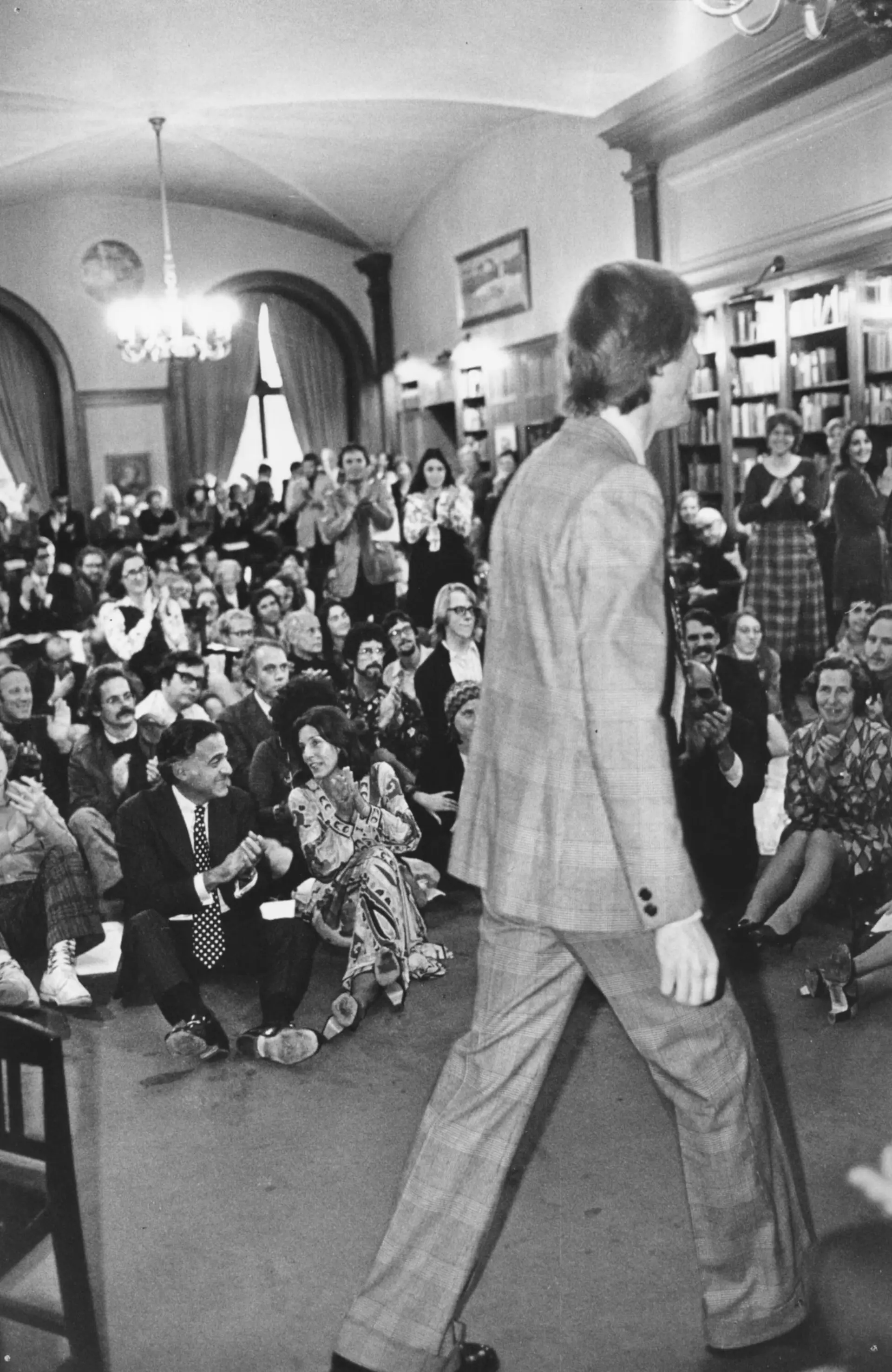 A man walks in front of an audience seated on the floor in a library