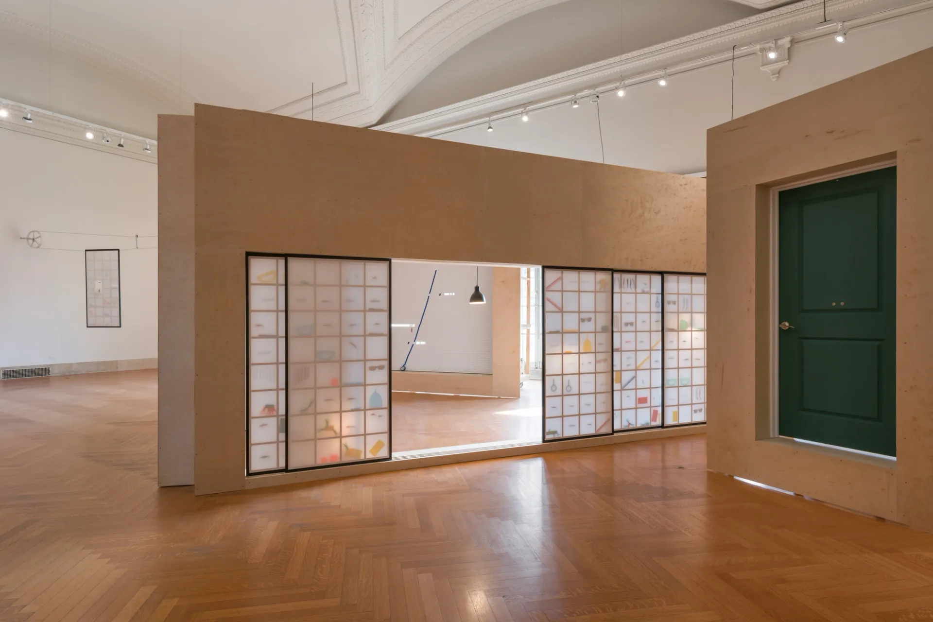An art installation with a row of siding paper doors with objects and a gridded wooden frame visible inside. The doors are partially opened at different degrees in a wooden wall positioned at an angle. Adjacent to it is a green door situated within a wooden panel.