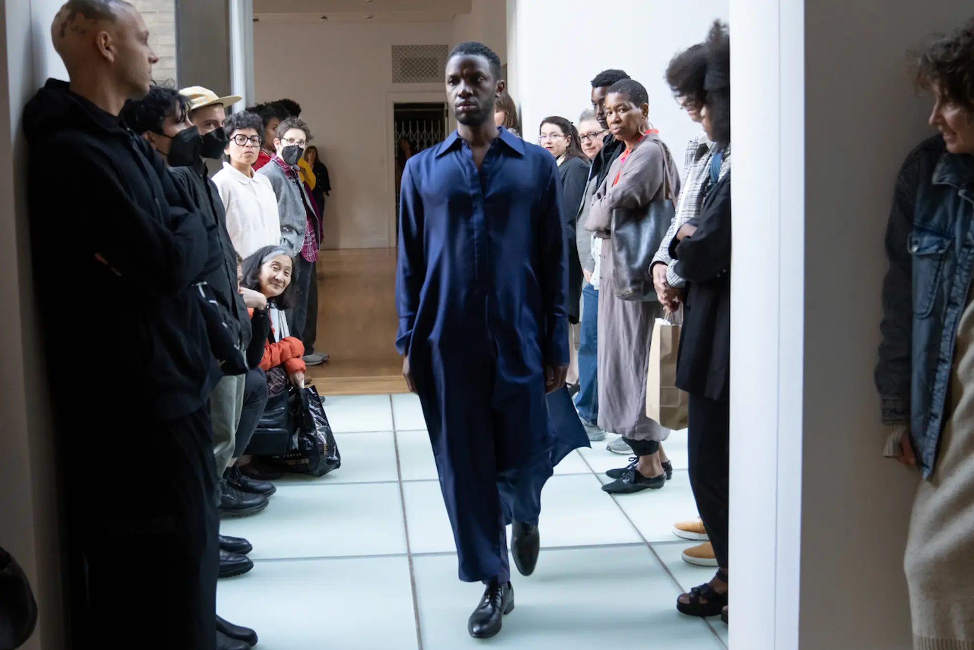 A young man in loose, lengthy clothing stands erect as he enters a space flanked by an audience of spectators on either side