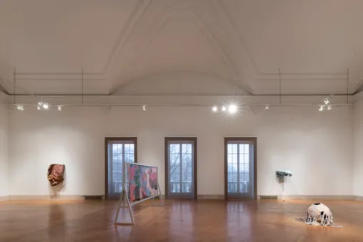 Various artworks are on display in a photograph of a gallery with three large windows at its center. An abstract sculpture in nude tones is mounted to a wall to the left of the windows while another sculpture extends from the walls to the right of the windows. In the foreground, a colorful painting mounted in a wooden armature and a round sculpture both stand on the floor. 
