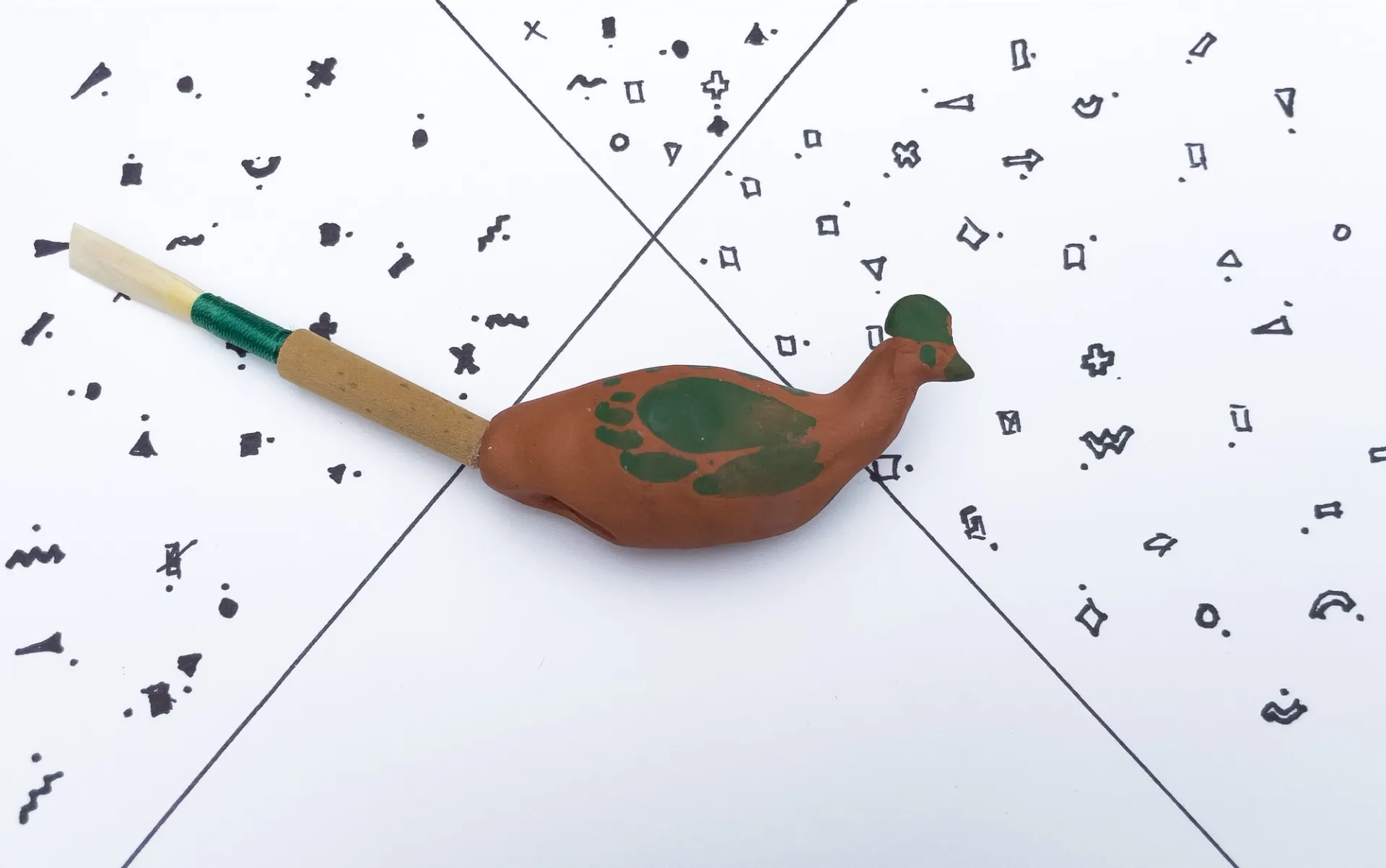 A clay bird with green painted feathers sits on a pattern on hand-drawn geometric shapes.  A clarinet reed protrudes from the back of the bird. 