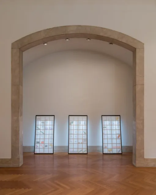 Three paper screens lean against a gallery wall inside an arched stone entryway. They are lit from behind and colorful objects in wooden grids are visible inside through the paper.