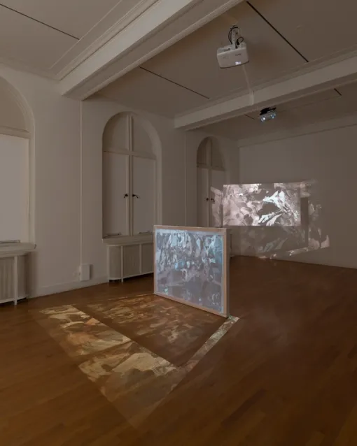 A translucent projection screen with a wooden frame rests on the floor of a darkened gallery. The projected image passes through the screen and expands on the floor. Just behind this screen another projection is visible on the wall.