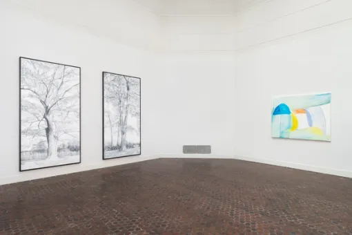 On a wall to the left, two large black and white paintings of trees hang beside one another. To the right, a smaller colorful painting hangs on the adjacent wall.