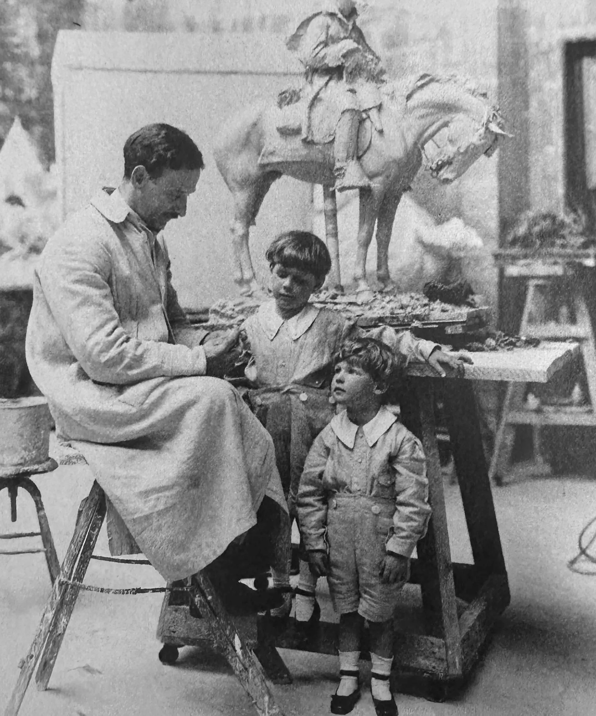 A black and white photograph shows a man in a white work coat sitting on a stepladder and looking down to his two sons beside him. Behind these figures, is a sculpture of a man on a horse standing on a workbench.
