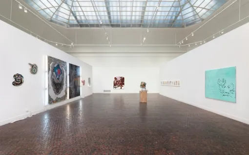A long gallery space displays small and large abstract artworks on the walls. Directly ahead, a sculpture sits on a wooden base.