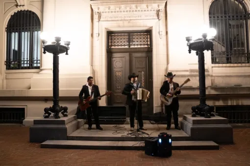 Outdoors at night, a band of three musical performers serenade an audience from the entryway steps of a building. To the right and left, band members perform on the guitar, and in the middle, a performer is on the microphone and accordion