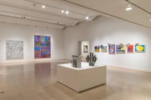 Two large paintings, one gray and the other an assortment of colors, hang mounted on the wall to the left. On the right, six colorful artworks of varying sizes hang beside one another. In the center of the room, three black and white sculptures sit on a large white pedestal.