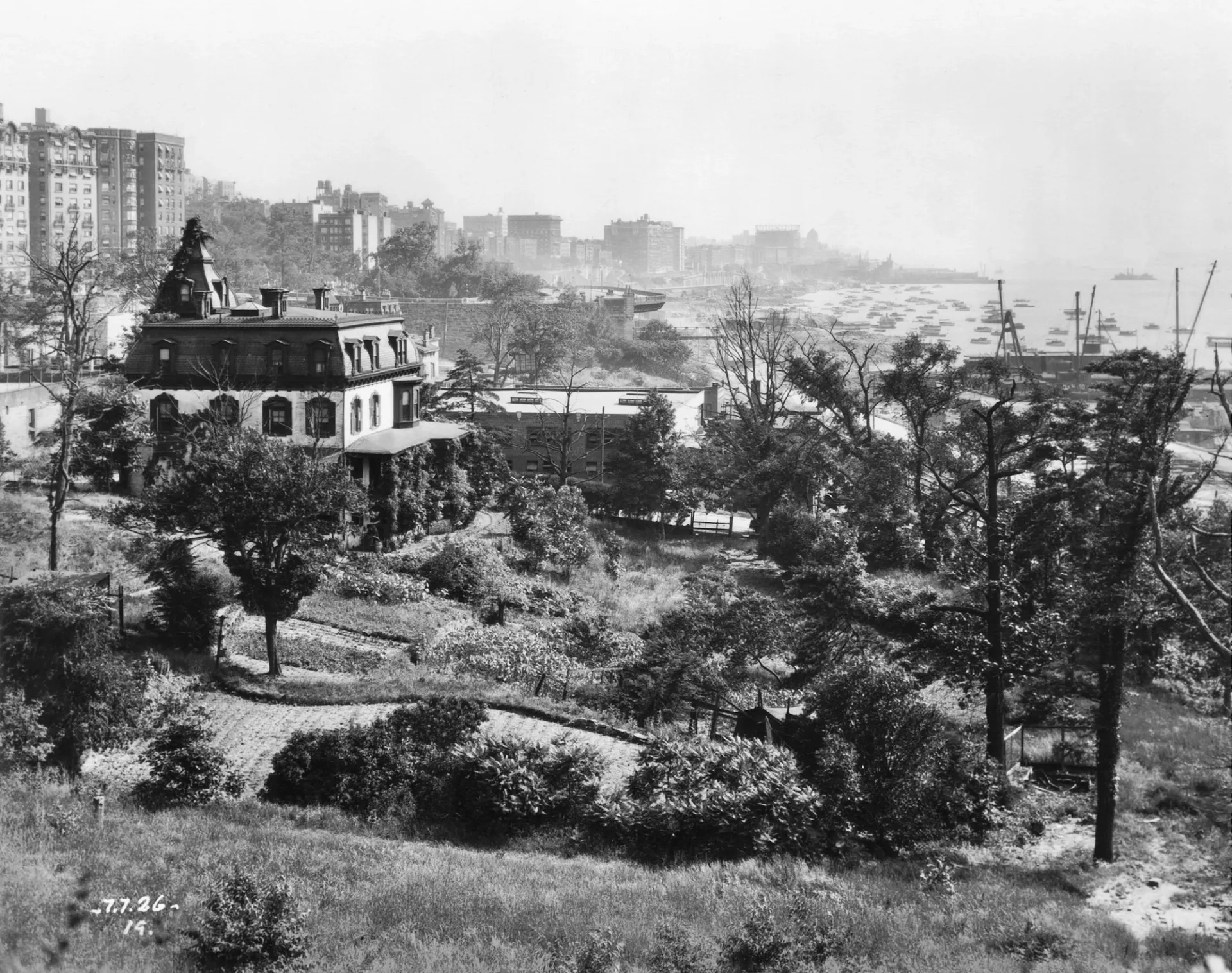 A three-story house surrounded by a lawn and gardens overlooks the Hudson River. Tall apartment buildings are visible in the background.