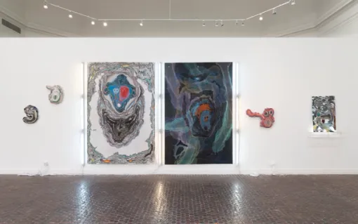 Two large works on paper with contrasting colors hang parallel to each other with a set of three white fluorescent lights set beside them. These two works are flanked by smaller mounted sculptures.
