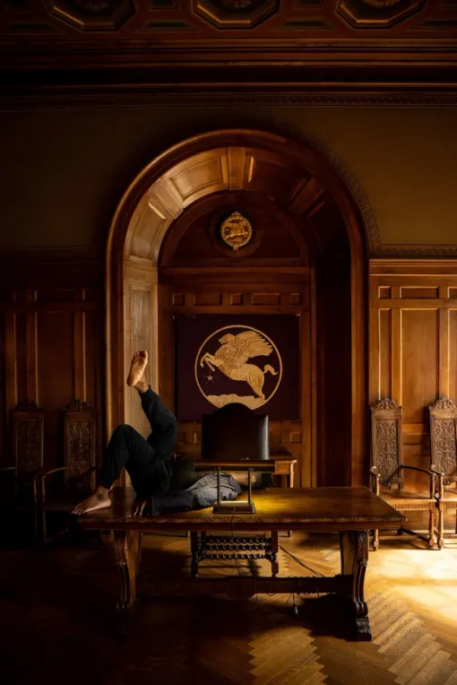 A performer reclines, one foot in the air, on a desk in a wood-paneled room