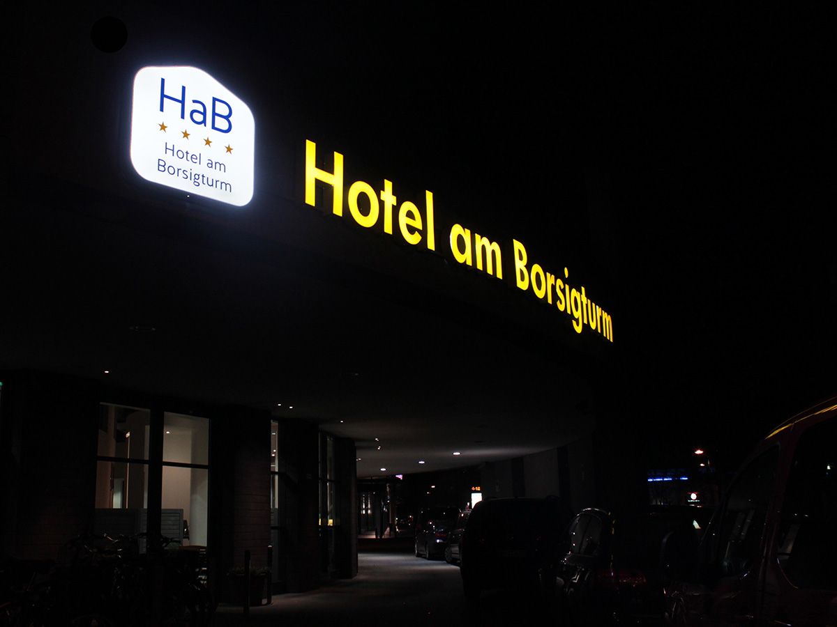 Gelbe Leuchtschrift „Hotel am Borsigturm“ und beleuchtetes Logoschild an der Hotelfassade bei Nacht