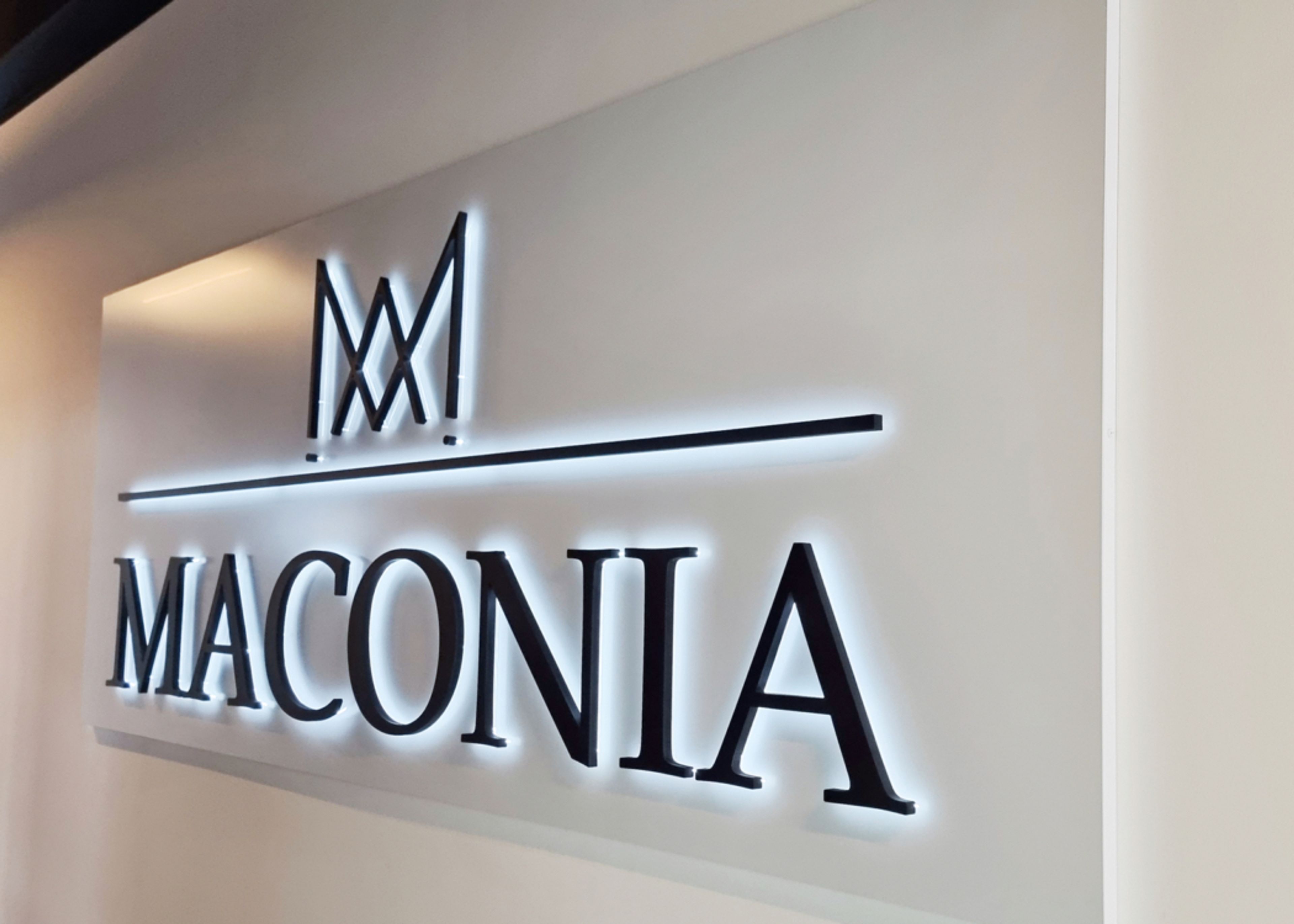 Logo und Schriftzug „Maconia“ aus dunklen Buchstaben mit hellem Lichtschein auf einer weißen Wandfläche, von der Seite