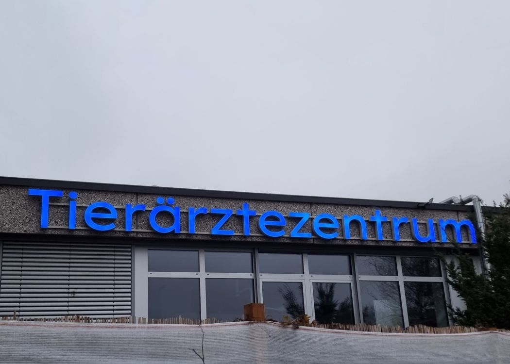 Schriftzug „Tierärztezentrum“ aus blauen Leuchtbuchstaben an der Gebäudefront, von schräg unten bei bewölktem Himmel