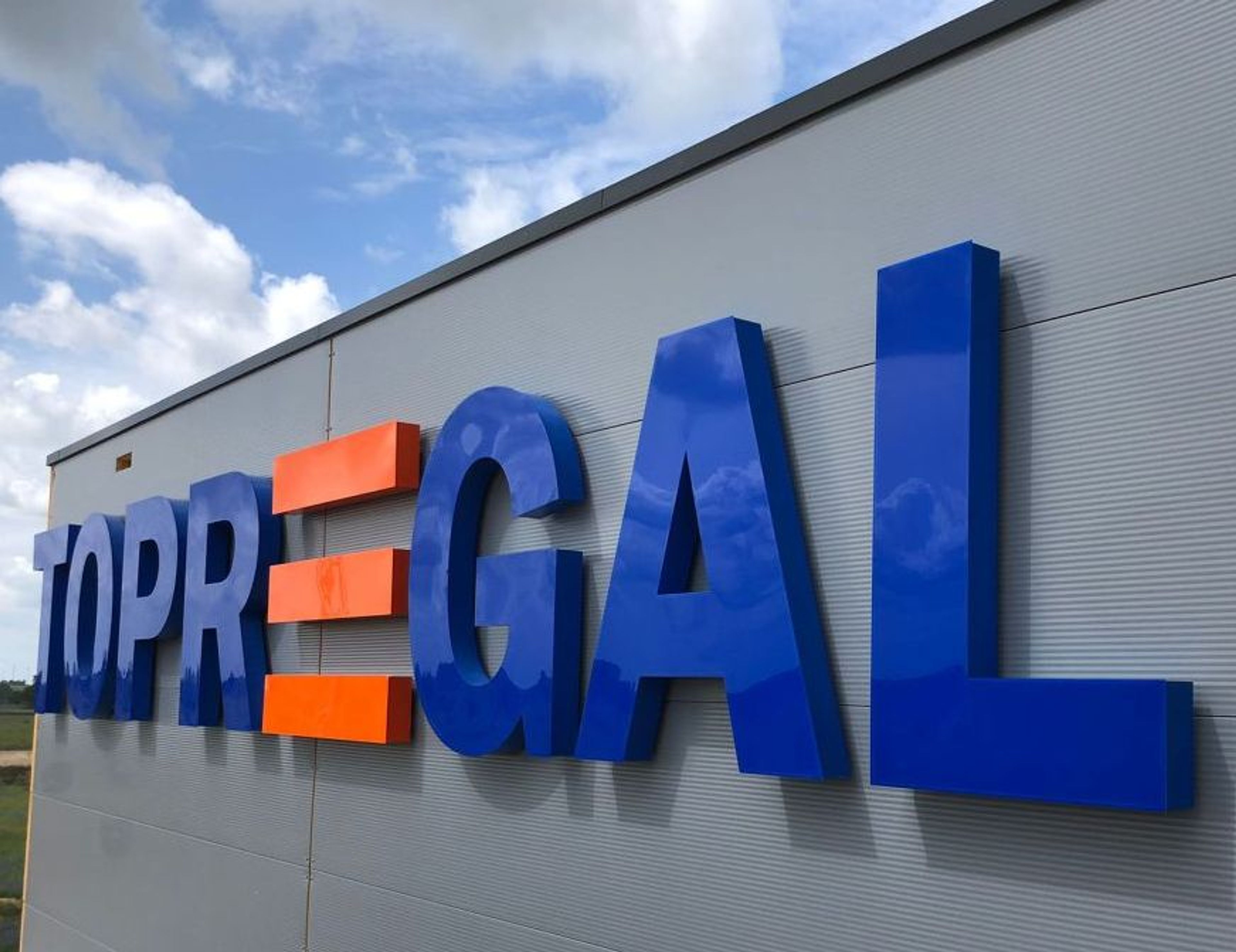 Große blaue Buchstaben mit orangefarbenen Balken an der Metallfassade einer Halle, von schräg unten bei Tag