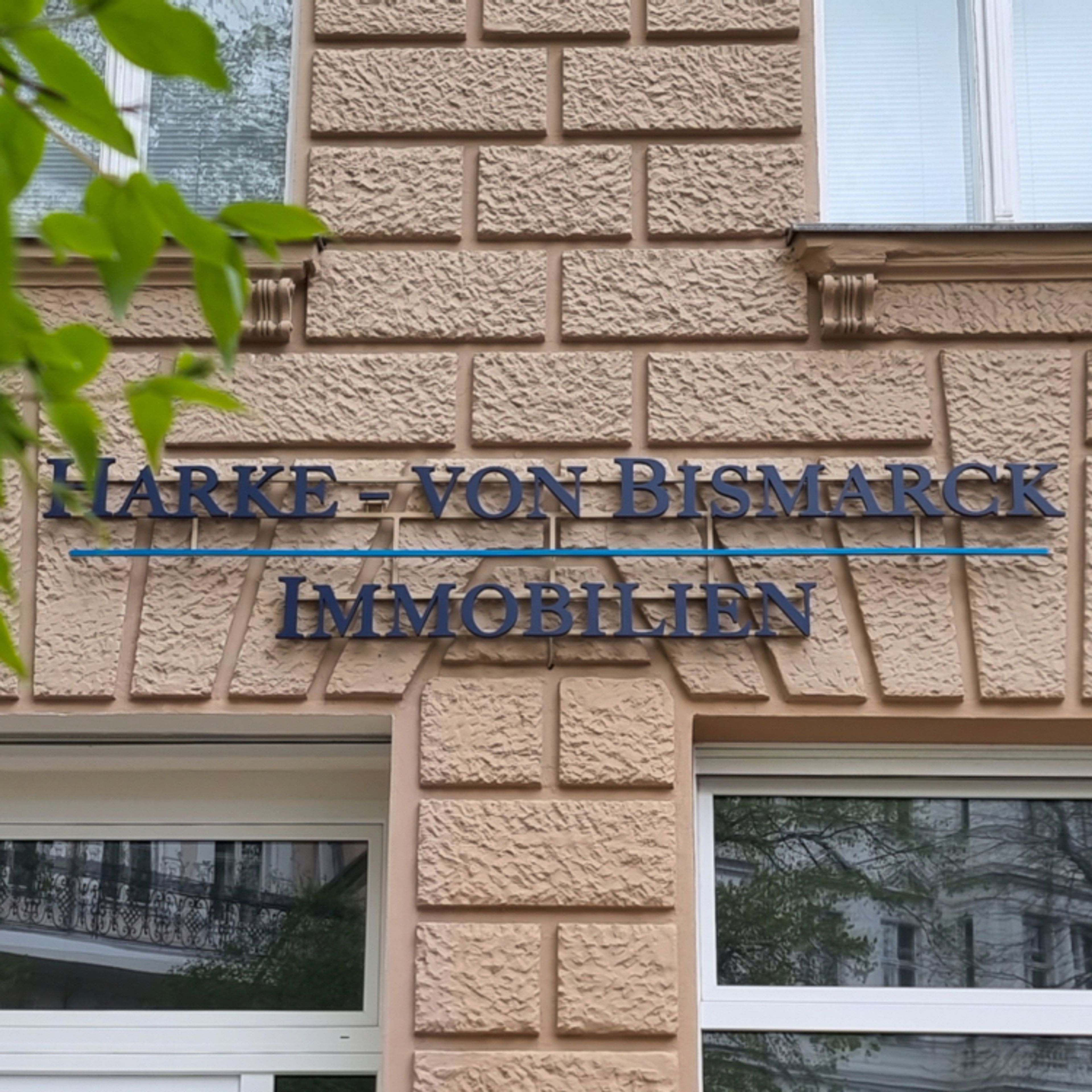 Schriftzug in zwei Zeilen an einer hellen Altbaufassade zwischen den Fenstern
