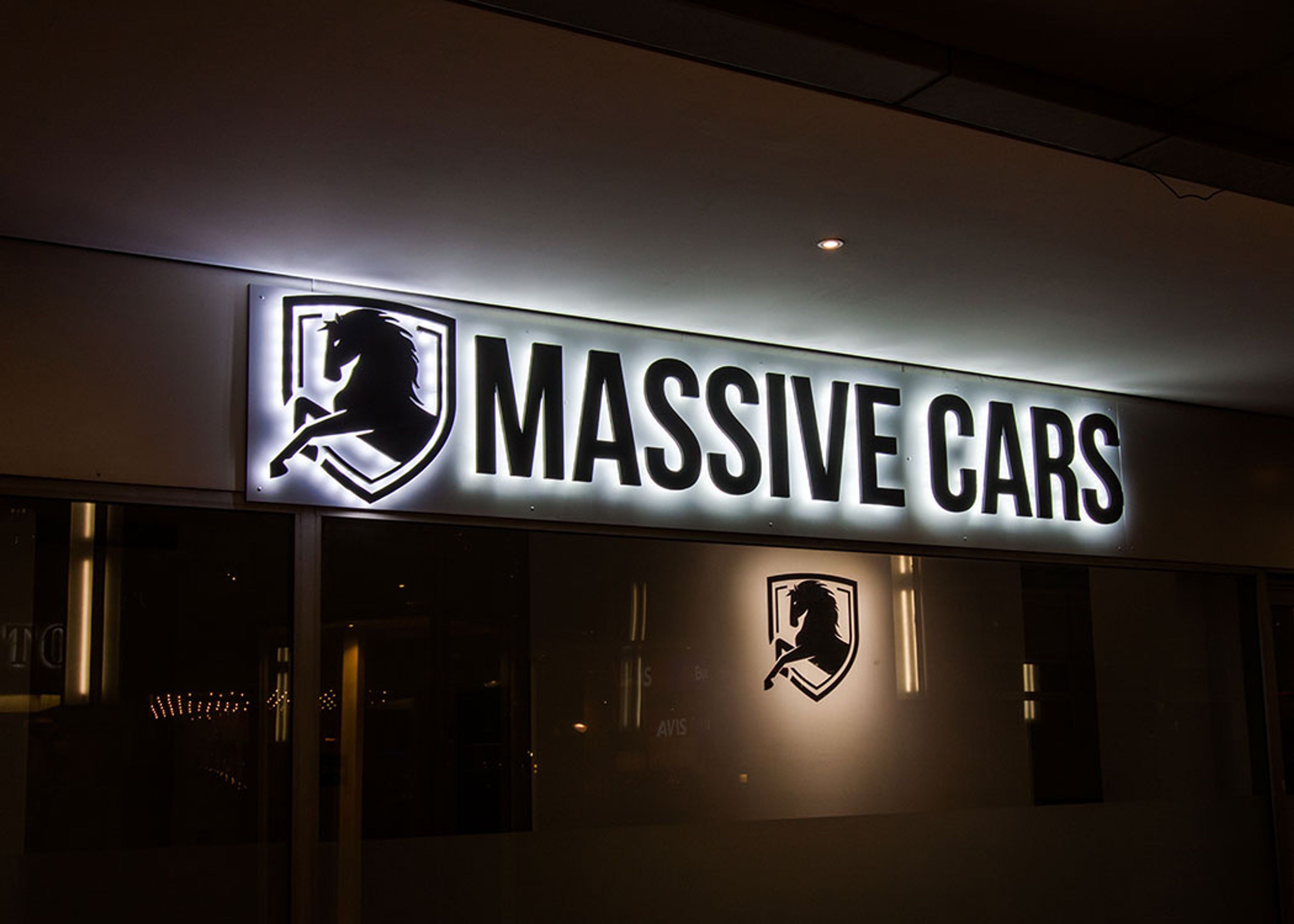 Buchstaben „Massive Cars“ und Pferdelogo mit hellem Lichtschein auf einer Platte über einem Schaufenster am Abend