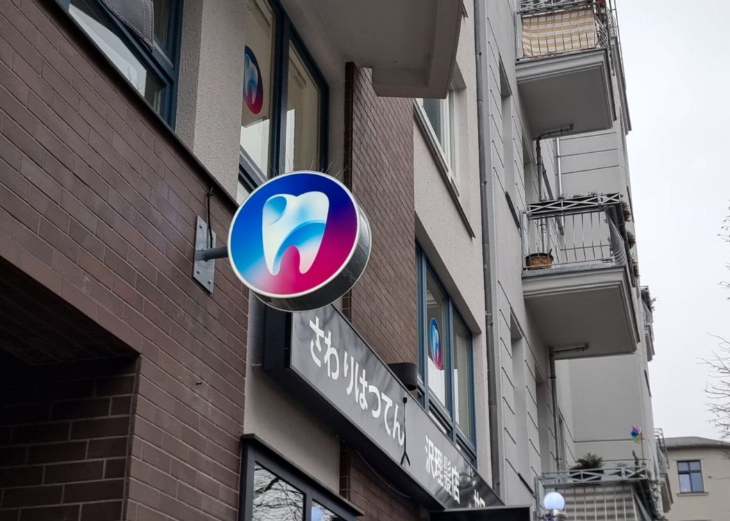 Runder Leuchtkasten mit Zahn-Logo quer zur Fassade eines Wohn- und Geschäftshauses, von schräg unten bei Tag