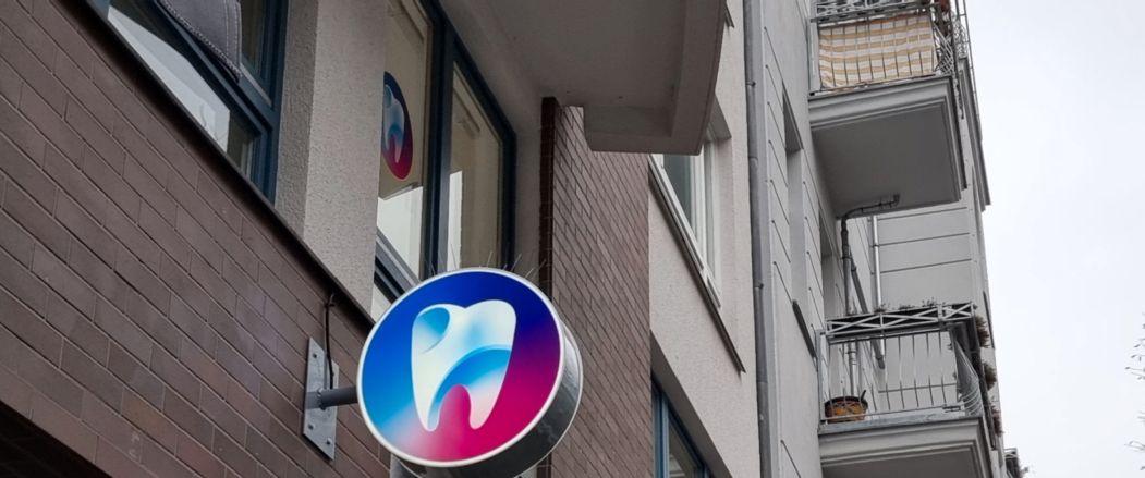 Runder Leuchtkasten mit Zahn-Logo quer zur Fassade eines Wohn- und Geschäftshauses, von schräg unten bei Tag
