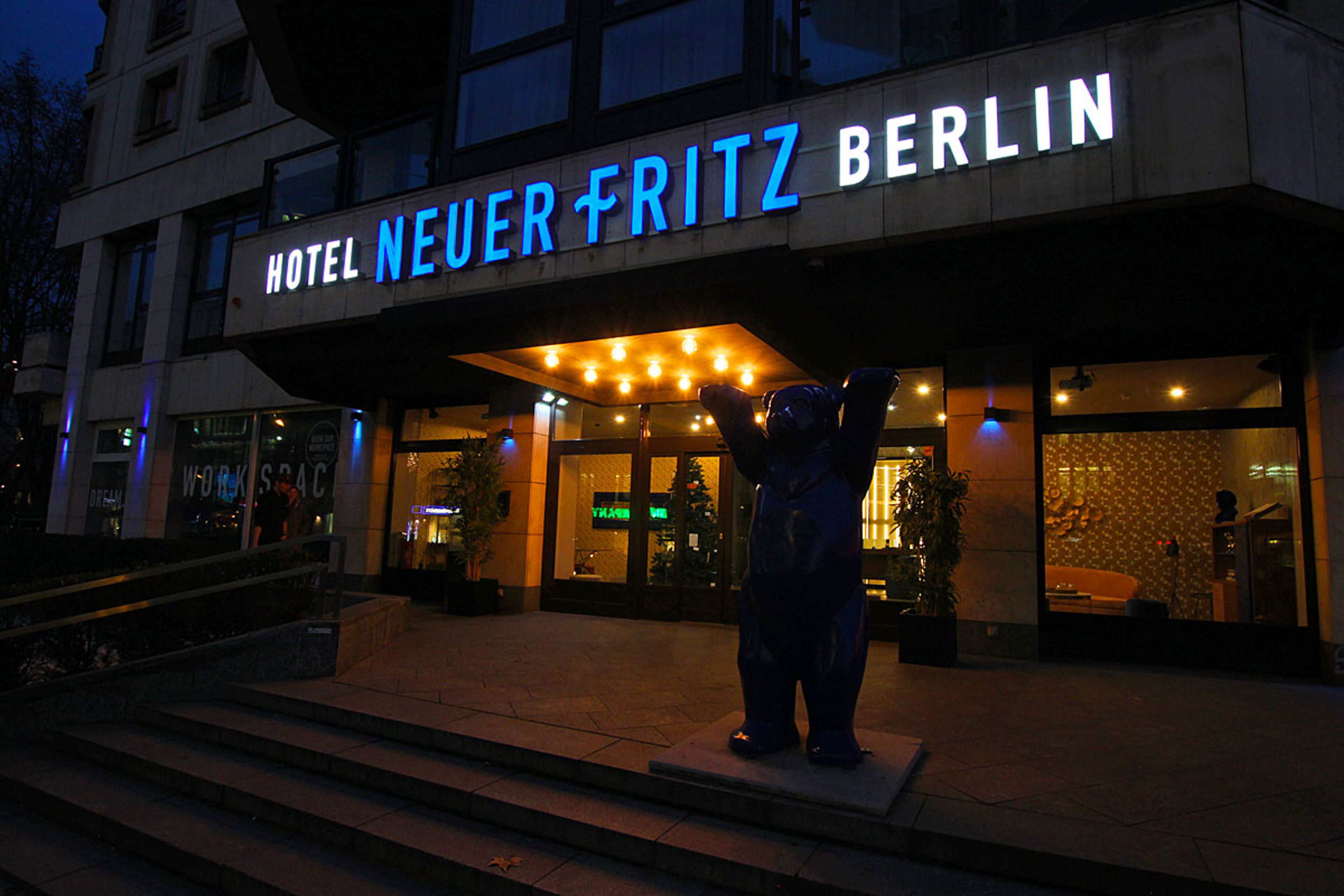 Leuchtschriftzug „Hotel Neuer Fritz Berlin“ über dem Hoteleingang am Abend, davor eine Bärenfigur auf der Treppe