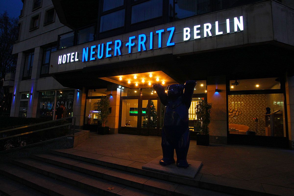 Leuchtschriftzug „Hotel Neuer Fritz Berlin“ über dem Hoteleingang am Abend, davor eine Bärenfigur auf der Treppe