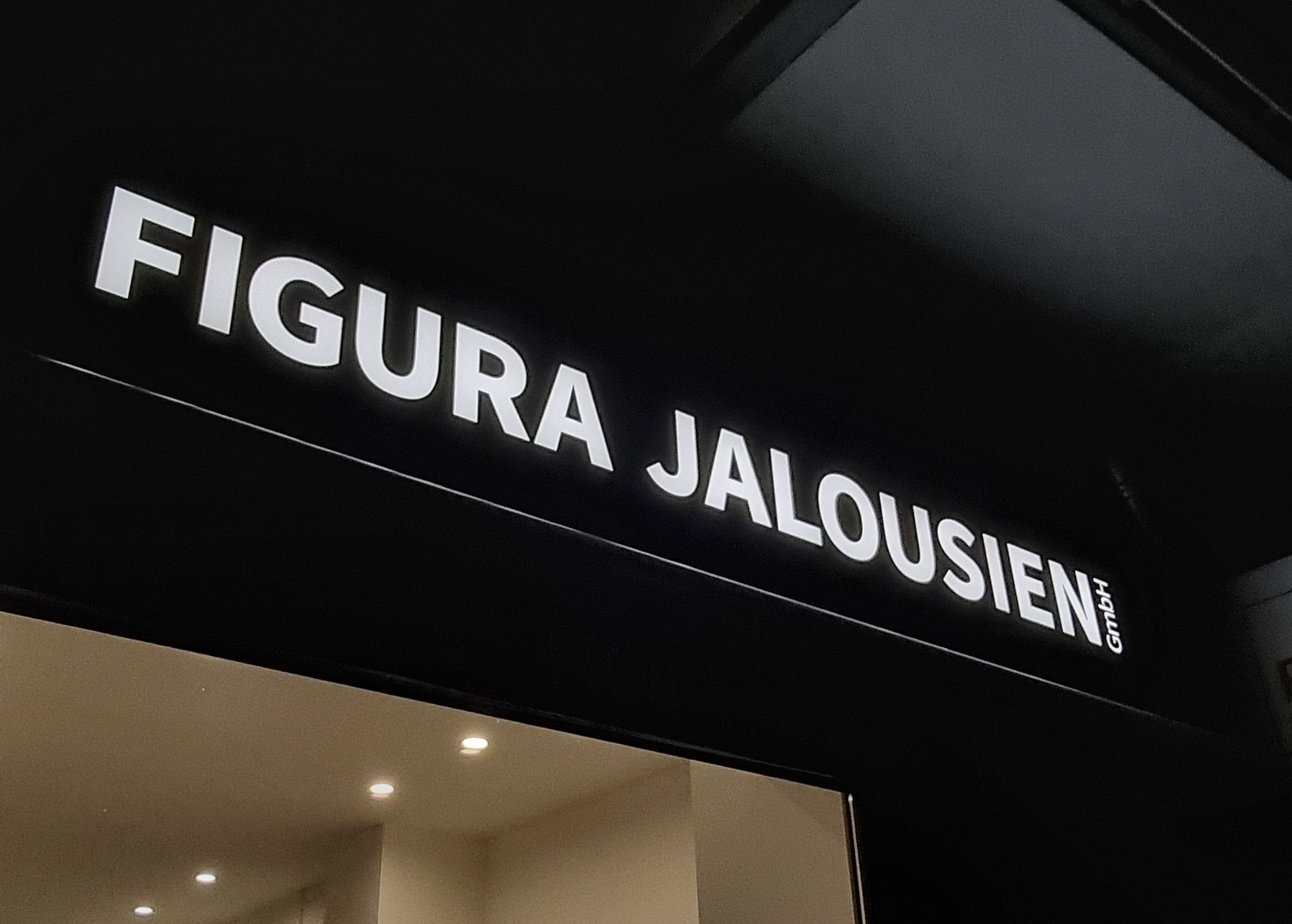 Weiß leuchtende Schrift „Figura Jalousien“ auf dunkler Blende über einem Schaufenster am Abend, Nahansicht