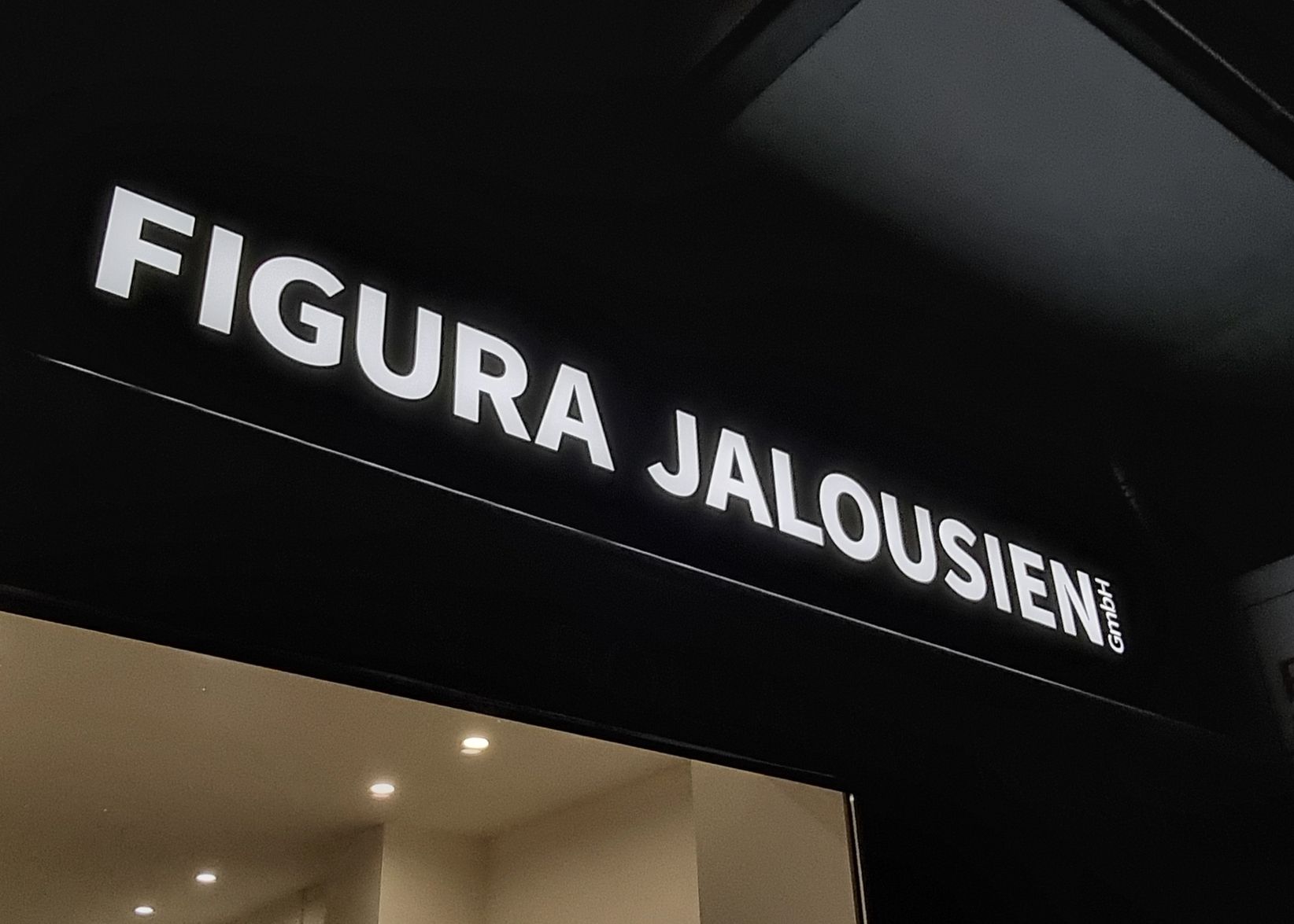 Weiß leuchtende Schrift „Figura Jalousien“ auf dunkler Blende über einem Schaufenster am Abend, Nahansicht