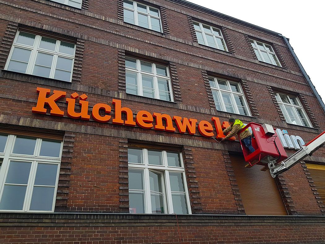 Monteur auf einer Hubarbeitsbühne bringt die orangefarbenen Buchstaben „Küchenwelt“ an der Backsteinfassade an