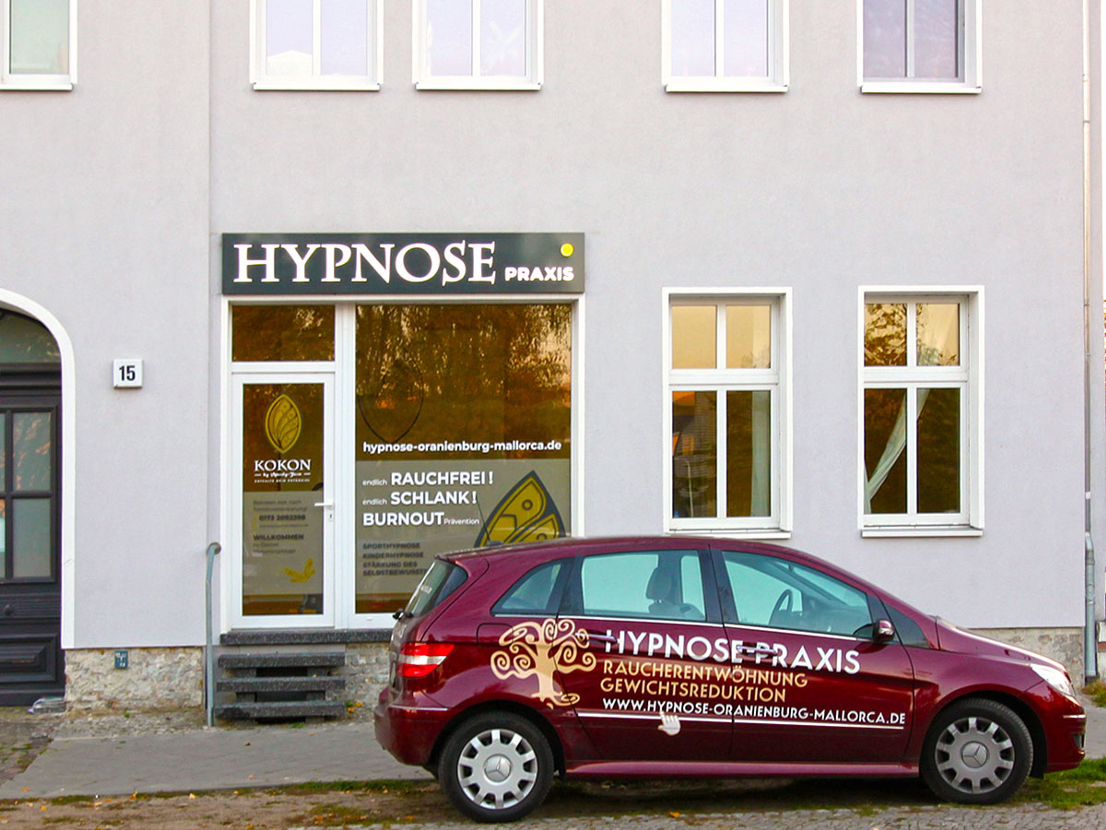 Leuchtkasten mit heller Schrift über Tür und Schaufenster einer Praxis, frontal bei Tag