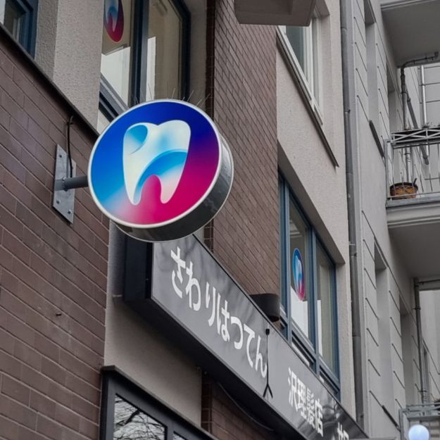 Runder Leuchtkasten mit Zahn-Logo quer zur Fassade, bei Tag