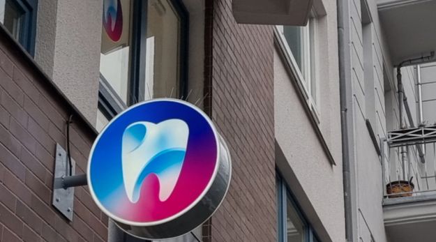 Runder Leuchtkasten mit Zahn-Logo quer zur Fassade, bei Tag