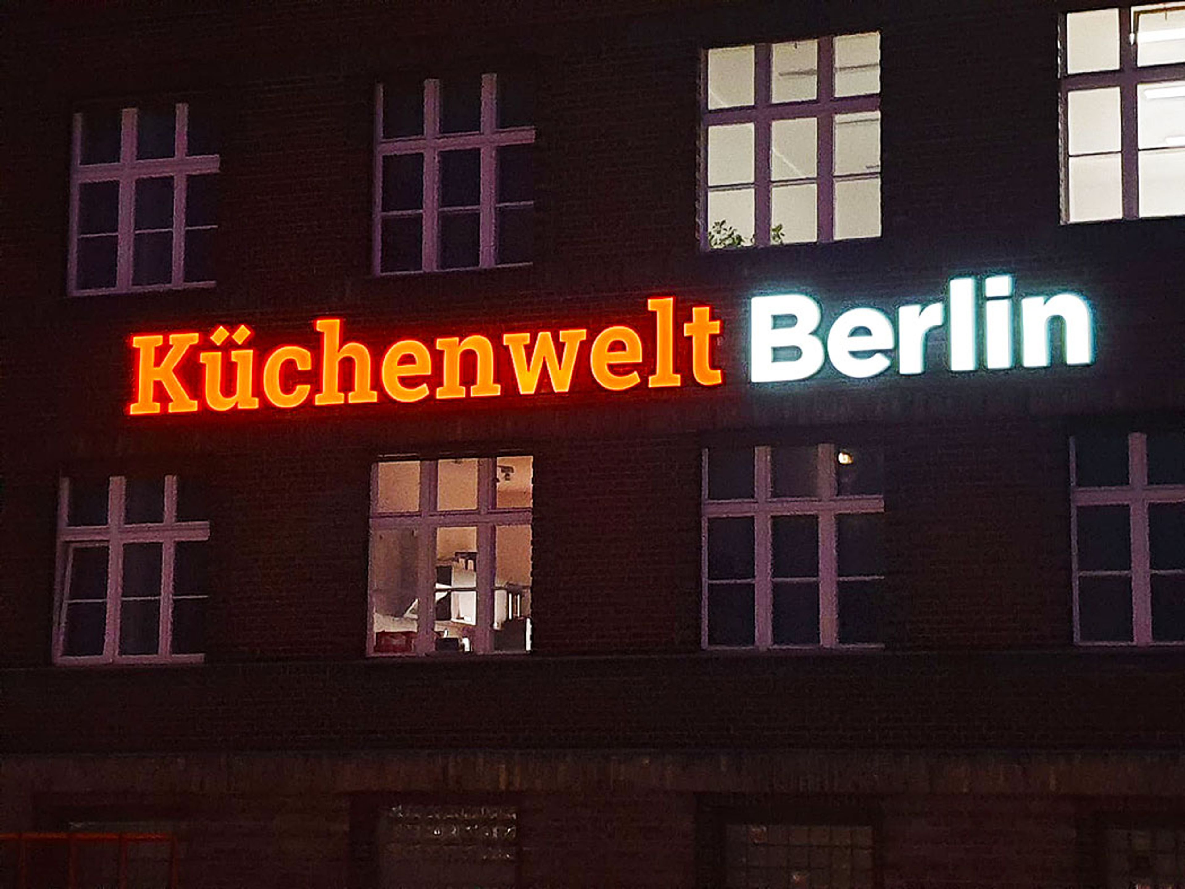 Mehrstöckige Backsteinfassade bei Nacht, zwischen den Fensterreihen leuchtet der Schriftzug „Küchenwelt Berlin“