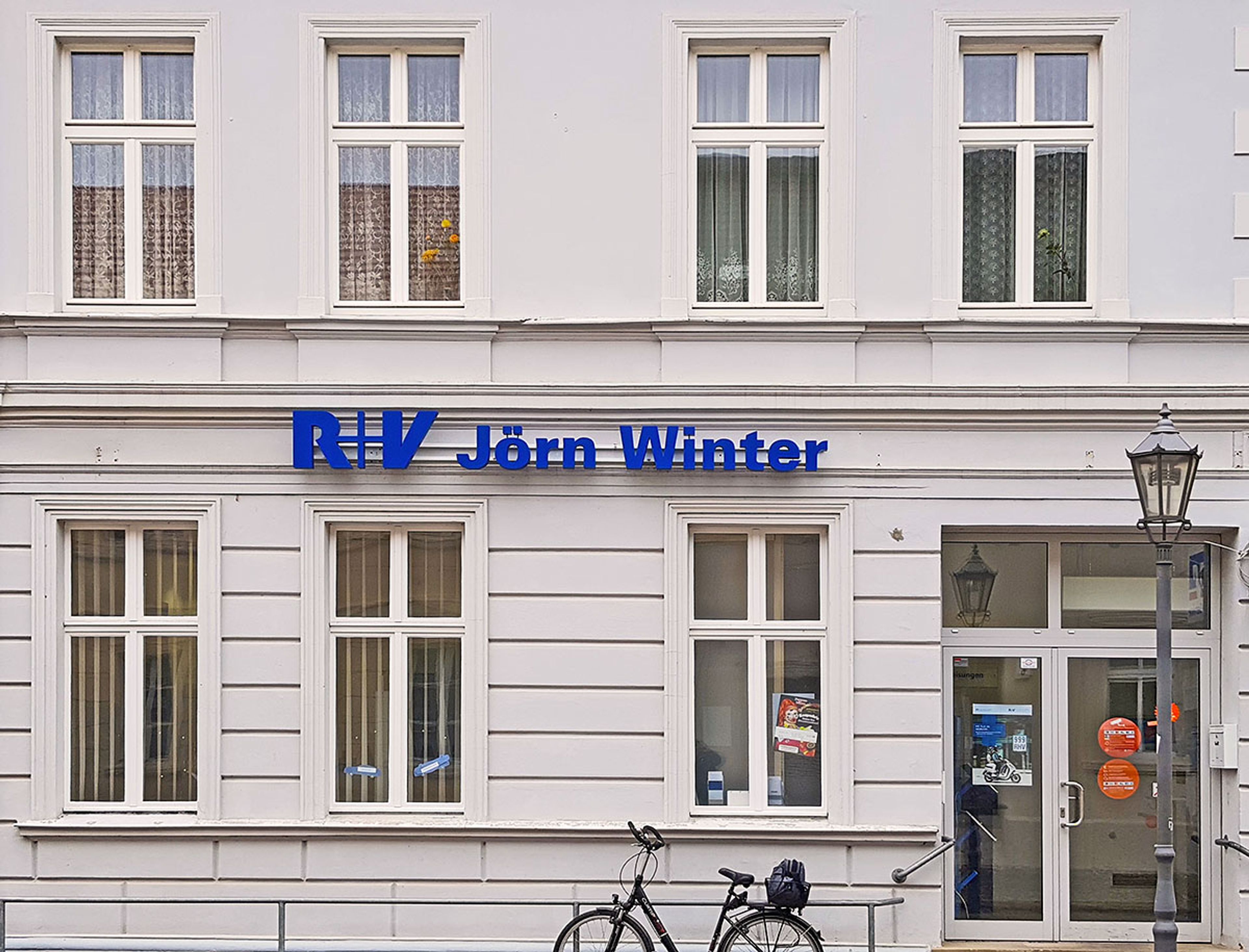 Helle Altbaufassade bei Tag, über den Fenstern im Erdgeschoss ein blauer Schriftzug eines Versicherungsbüros