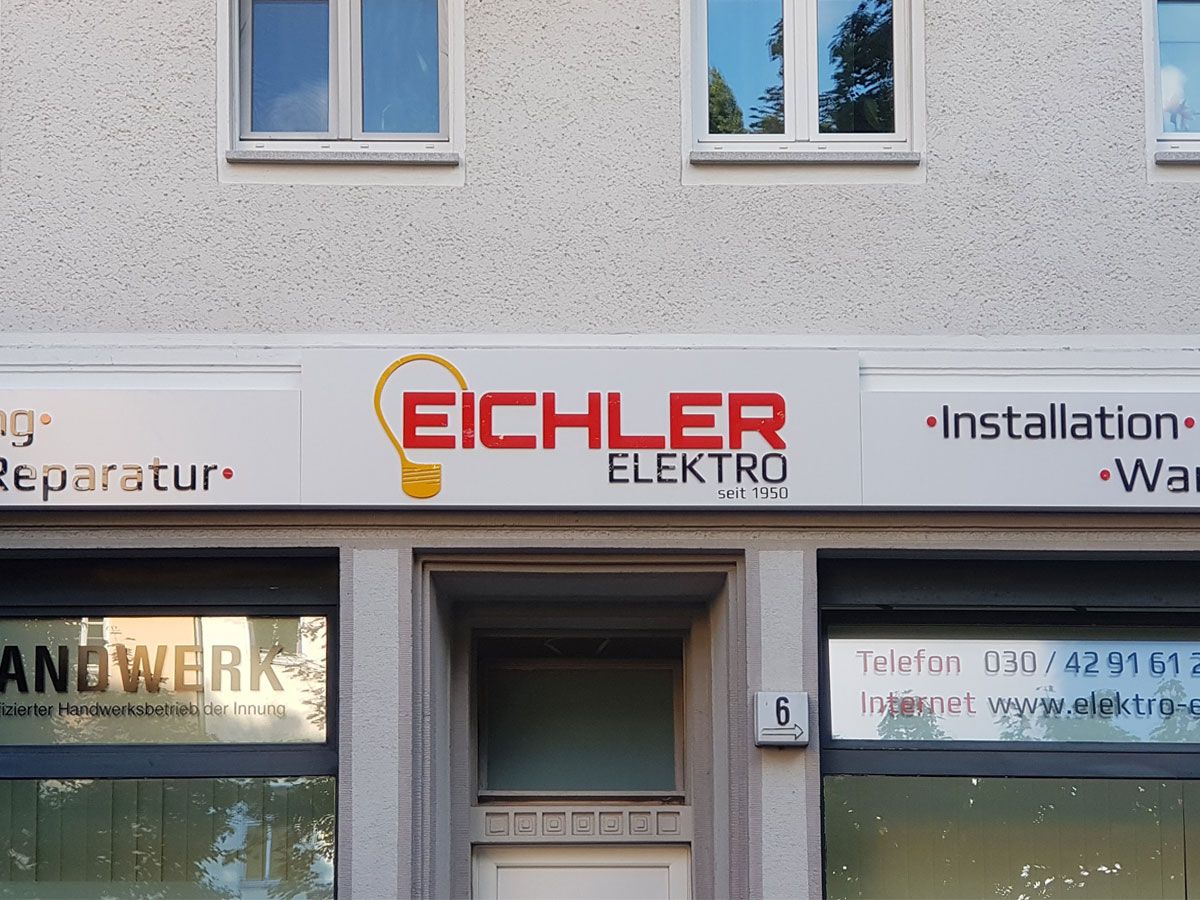 Weißer Leuchtkasten mit oranger und roter Schrift an einer Hausfassade, bei Tag