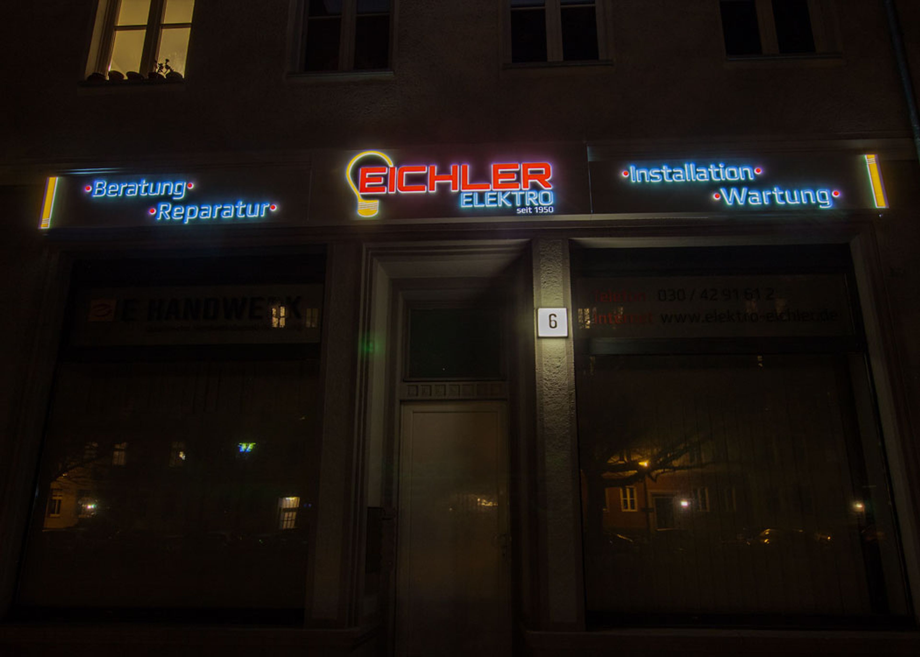 Ladenfront bei Nacht frontal, über Eingang und Schaufenstern leuchten Schriftzüge in Blau und Rot und ein Glühbirnen-Logo