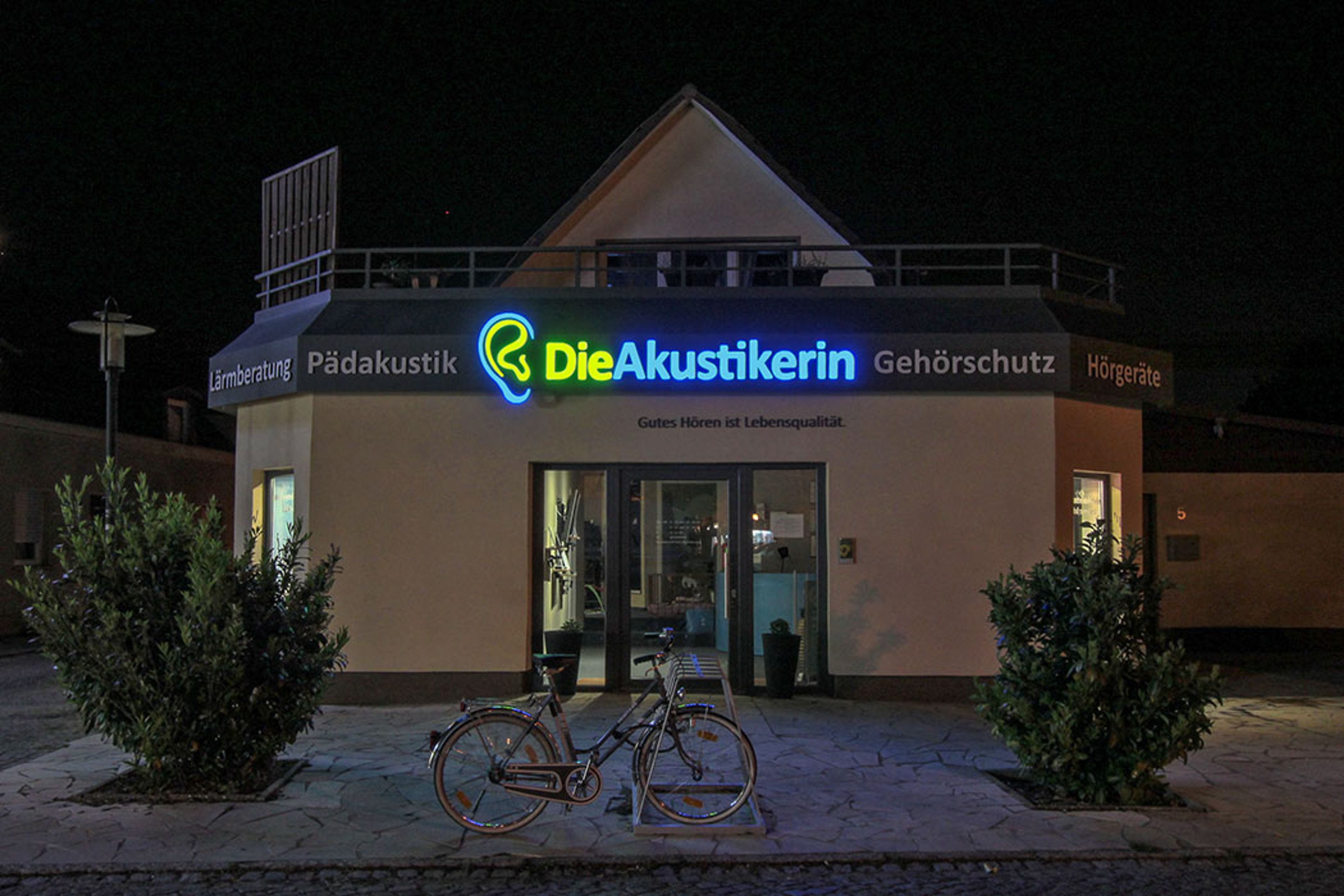 Ladengebäude bei Nacht aus etwas größerer Entfernung, in der Mitte der Blende leuchtet „Die Akustikerin“