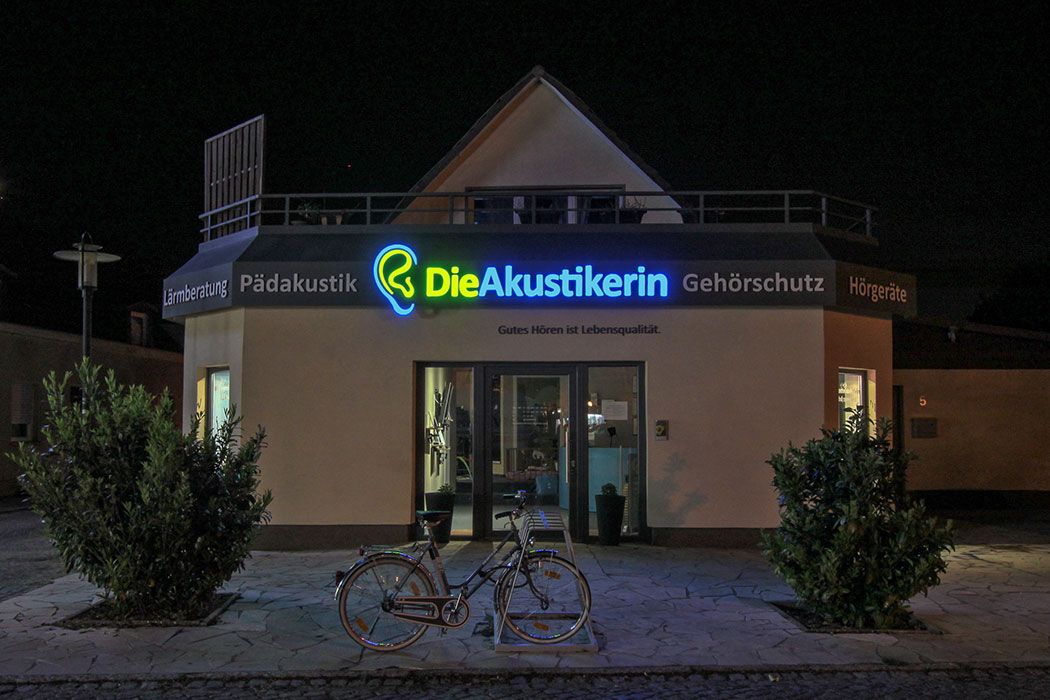 Ladengebäude bei Nacht aus etwas größerer Entfernung, in der Mitte der Blende leuchtet „Die Akustikerin“