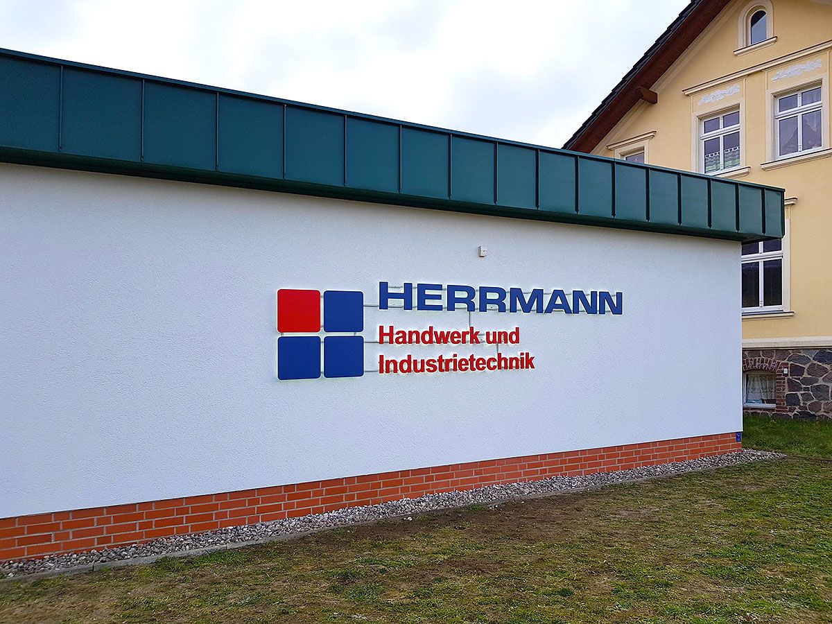 Flachbau mit weißer Putzfassade bei Tag, darauf ein Logo aus vier Quadraten und ein Schriftzug in Blau und Rot
