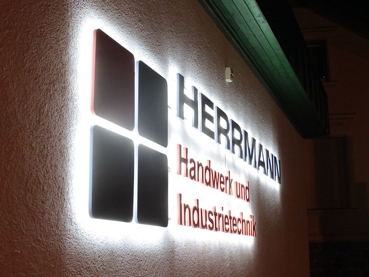 Logo aus vier Quadraten und Schriftzug mit hellem Lichtschein an einer Putzfassade bei Nacht, von schräg gesehen